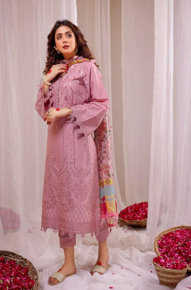 A woman in a pink Pakistani dress stands gracefully in front of a vibrant flower arrangement.