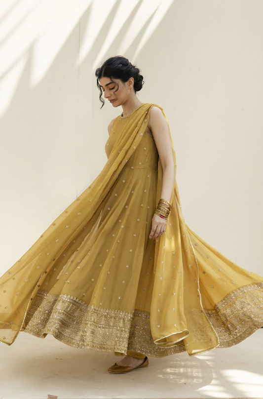 A woman in a yellow dress walks gracefully along a path, surrounded by greenery.