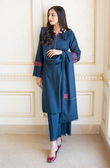 A woman in a blue Pakistani suit stands gracefully against a textured wall, showcasing her elegant attire.
